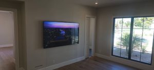 Modern living room featuring a flat-screen TV mounted on the wall, with large windows allowing natural light and showcasing a view of palm trees outside.